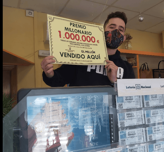 Hombre con mascarilla muestra cartel de premio millonario vendido en una administración de lotería.