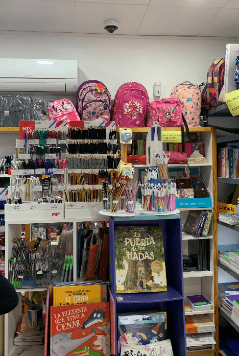 Estantes de papelería con mochilas rosas, diversos bolígrafos, lápices y libros infantiles de cuentos.